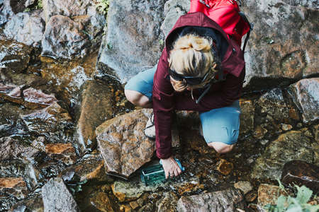 Woman taking pure water to bottle from mountain stream during trekking in mountains. Hiker crouching on rocks, filling bottle up with cold mountain water. Enjoying the outdoors in the summer trip vacationの写真素材