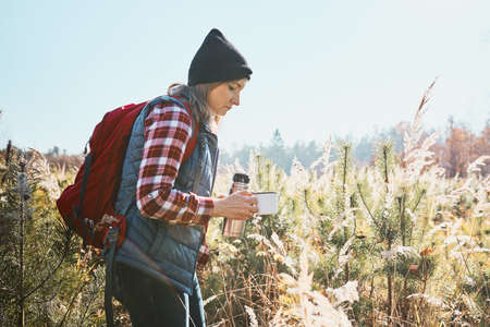 Woman taking break and relaxing in bright warm sunlight with cup of coffee during summer trip. Woman standing on trail and looking away. Woman with backpack hiking through tall grass along path in mountains. Spending summer vacation close to natureの写真素材
