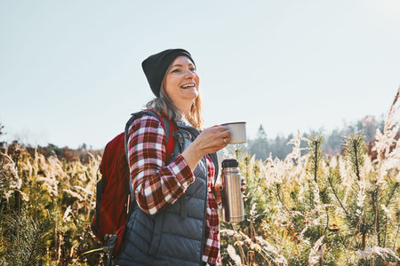 Smiling woman relaxing and enjoying the coffee during vacation trip. Woman standing on trail and...
