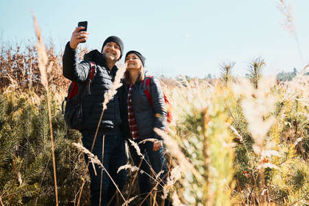 Hugging couple taking selfie while vacation trip. Hikers with backpacks on way to mountains. People walking through tall grass along path in meadow on sunny day. Active leisure time close to natureの写真素材