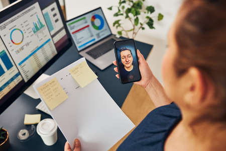 Businesswoman having video chat on mobile phone with her colleague. Businesswoman working with data on charts, graphs and diagrams on computer screen. Woman holding smartphone sitting at desk in officeの写真素材