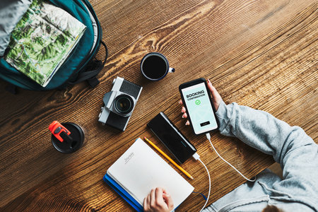 Woman planning next vacation trip. Booking hotel online using smartphone. Ordering service. Charging mobile phone with power bank. Using technology while travelling. Girl sitting at wooden table with camera and backpack during journey. Travel plan conceptの写真素材