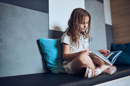 Schoolgirl reading book in school library. Primary school pupil is engaged in book. Child doing homework. Smart girl learning from book. Benefits of everyday reading. Child curiosityの写真素材