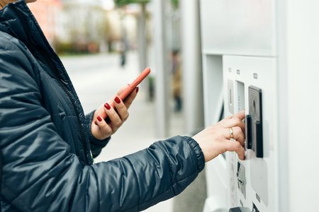 Woman buying ticket at ticket machine paying using mobile payment app on smartphone. Female buying...