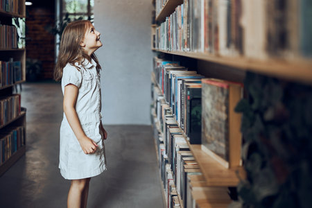 Schoolgirl looking at books on top bookshelf in school library. Smart girl selecting literature for reading. Books on shelves in bookstore. Learning from books. School education. Benefits of everyday reading. Child curiosity. Back to schoolの写真素材