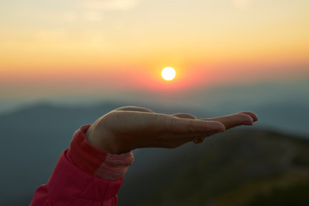 Sun over female hand. Sunrise in mountains. Ready for adventure. Natural mountain landscape with illuminated misty peaks, foggy slopes and valleys, blue sky with orange yellow sunlightの写真素材