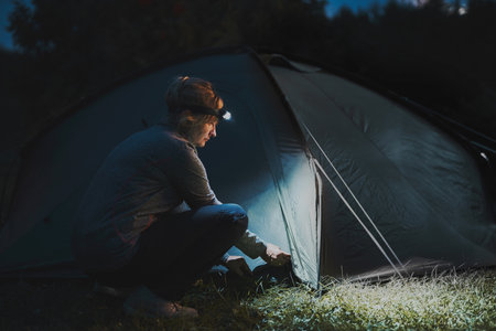 Summer vacation. Woman opening tent at camping in the evening using head lamp. Summer trip. Preparing campsite. Spending vacation outdoors close to natureの写真素材