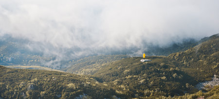 Vacation in mountains. Woman standing on rock on top of hill looking at coming fog. Hiker wearing yellow jacket. Active woman hiking in mountains. Foggy mountain landscapeの写真素材