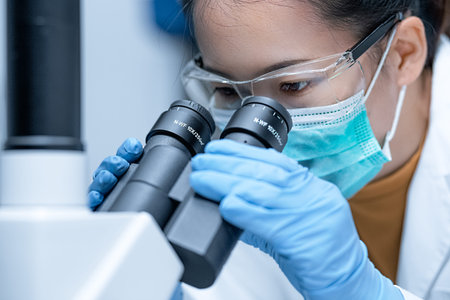 Closeup shot of a female researcher wearing safety goggles looking with the inverted microscope to...