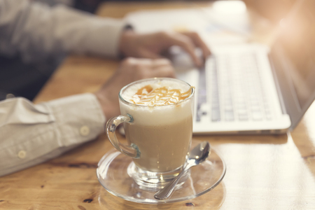 man's hand working with laptop computer with cup of hot latte coffee on wooden table, selective focus and vintage toneの写真素材