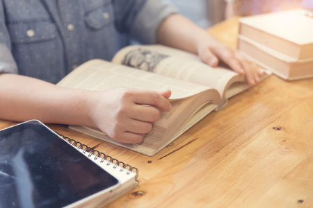 woman's hand reading book with pen, tablet and notebook on wooden table, selective focus and vintage...