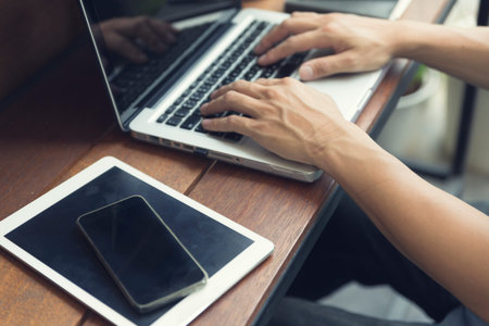 man's hand typing on laptop computer with smartphone and digital tablet on wooden table, selective...