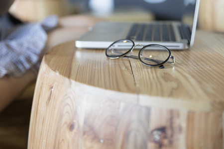 eyeglasses with laptop computer notebook on wooden desk with background of woman sitting on chairの写真素材