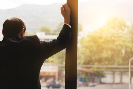 back of businessman in suit looking out through office windowの写真素材