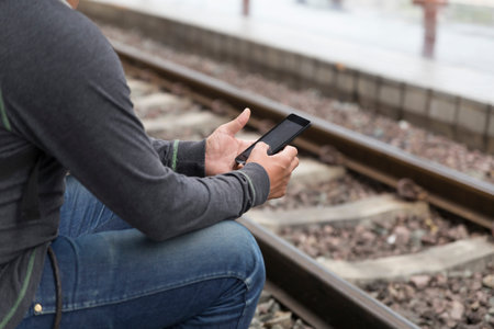 young man holding mobile phone sitting on platform at train station - travel conceptの写真素材