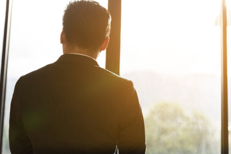 back of businessman in suit looking out through office windowの写真素材