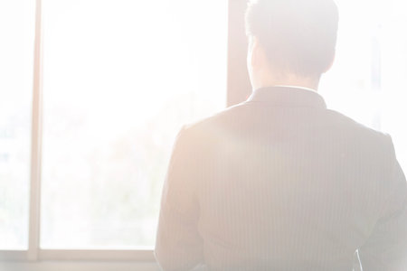 back of businessman in suit looking out through office window with flare