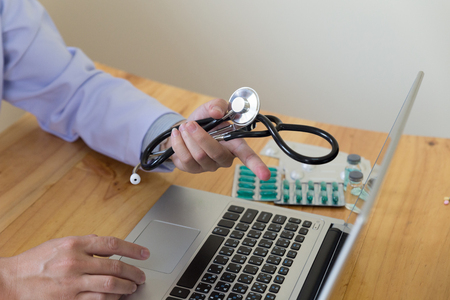 Male doctor, physician in white coat holding stethoscope pointing at computer screen showing something in medical office clinic with drug on deskの写真素材