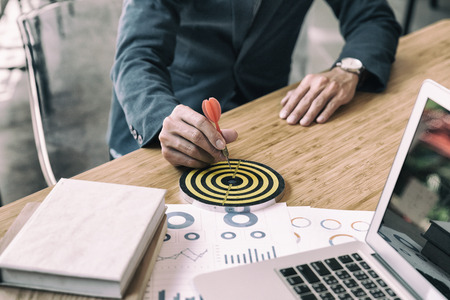 businessman gesturing while holding dart attached to target on dartboard on desk in office. Business, aim, achievement and success concept.の写真素材