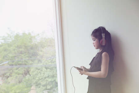 Pretty asian girl holding smart phone and listening to music with her headphones near window in roomの写真素材