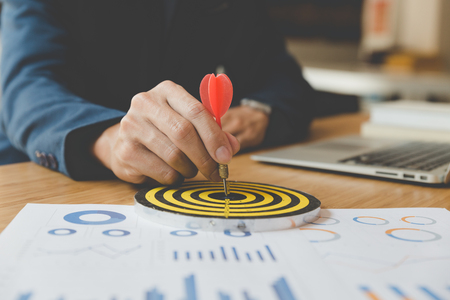 businessman gesturing while holding dart attached to target on dartboard on desk in office. Business, aim, achievement and success concept.の写真素材