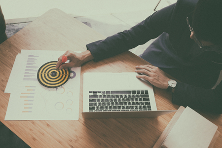 businessman gesturing while holding dart attached to target on dartboard on desk in office. Business, aim, achievement and success concept.の写真素材