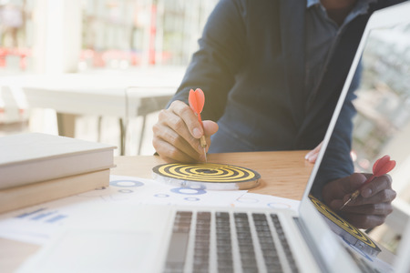 businessman gesturing while holding dart attached to target on dartboard on desk in office. Business, aim, achievement and success concept.の写真素材