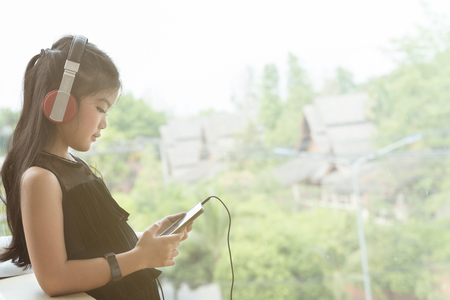 Pretty asian girl holding smart phone and listening to music with her headphones near window in roomの写真素材