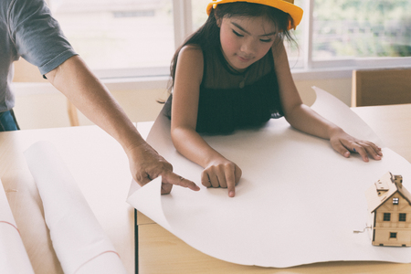asian little girl in protective construction helmet studying, learning engineering and architecture on blueprint with guidance of her teacher - childhood education conceptの写真素材