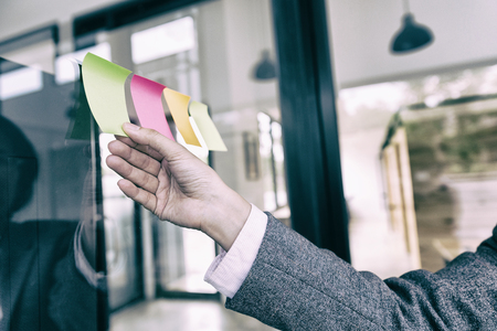 Female business person hand holding blank sticky paper note on glass wall in officeの写真素材