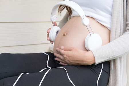 Young asian woman sitting and relaxing on balcony with headphones on her belly playing music to a baby in the womb.  Beautiful female smiling with pleasure expecting child having rest in front of her houseの写真素材