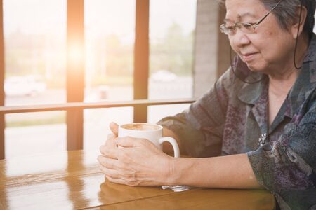 asian elderly woman sitting and resting in coffee shop with cup of cappuccino coffee. Elder senior lady in cafeの写真素材