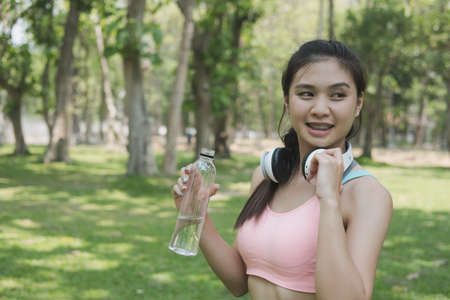 young beautiful asian fitness athlete woman holding drinking water after work out exercising at summer green park. Concept of healthy lifestyleの写真素材
