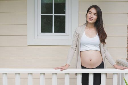 Young asian woman standing and relaxing on balcony smiling with pleasure. Beautiful female expecting child having rest in front of her houseの写真素材