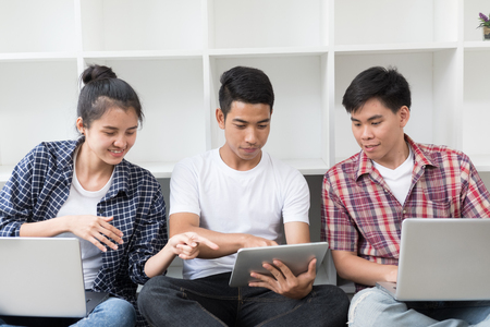 young asian people are using different gadgets and smiling, sitting near white wall. Male and female students studying using laptop computer, tablet. Education social media concept.の写真素材