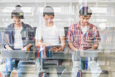 young asian people are using different gadgets and smiling, sitting near white wall. Male and female students studying using laptop computer, tablet. Education social media concept double exposure with libraryの写真素材
