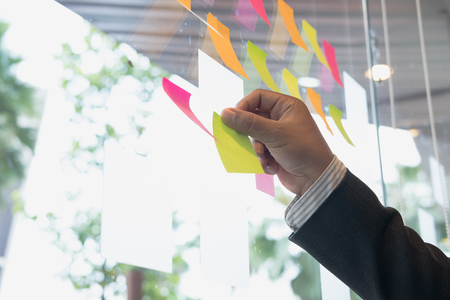 Businessman look at the adhesive notes on glass wall in meeting room. Sticky note paper reminder schedule for discussing idea in office. business, teamwork, brainstorming conceptの写真素材