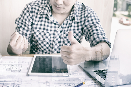 young male engineer worker raise his fist feeling happy and successful. Architect man show thumb up gesture with gladness after achieve construction project. Business and people concept double exposure with craneの写真素材