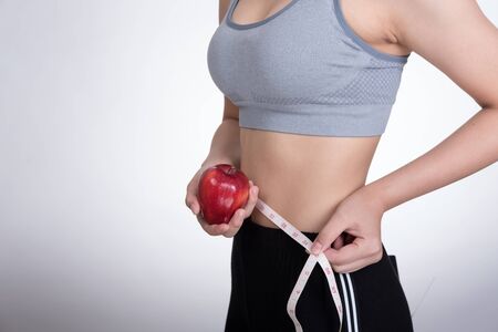 beautiful sporty fitness woman use measuring tape to measure the waist. young girl with red apple taking measurements of her body against white background. diet, sport and health conceptの写真素材
