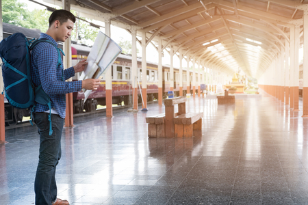 young asian man with backpack standing on platform at train station. backpacker or traveler look at map while waiting for train. journey, trip, travel conceptの写真素材
