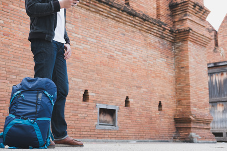 young traveler, handsome asian man wearing black jacket and blue jeans standing near old orange brick wall with mobile smart phone and backpackの写真素材