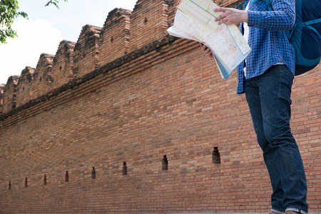 young traveler reading map. handsome asian man wearing blue shirt and jeans standing near old orange brick wall with backpackの写真素材