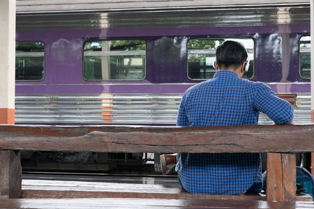 young asian man with backpack sitting on platform at train station. backpacker or traveler with headphone listening to music from mobile phone while waiting for train. journey, trip, travel conceptの写真素材