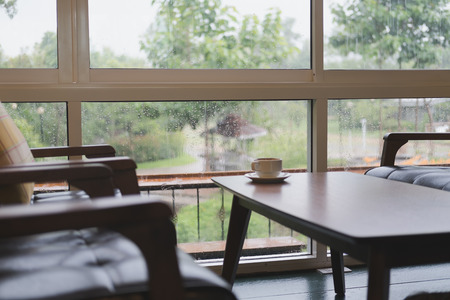 coffee cup on wood table, chair and brown leather sofa near window with garden view at restaurant or cafe in rainy dayの写真素材