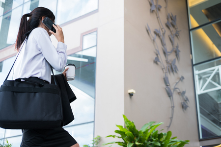 happy businesswoman holding mobile phone & coffee outside office building. beautiful young asian woman with tea go to work outdoors. female adult with disposable paper cup walking in business areaの写真素材
