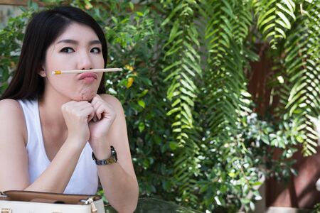 attractive asian woman sit at outdoor cafe. young female adult with natural make-up put pencil on mouth at coffee shop. happy teenager make funny faceの写真素材