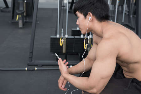 young man use mobile phone in fitness center. male athlete listen to music in cellphone in gym. sporty asian guy resting in health club after working out.の写真素材