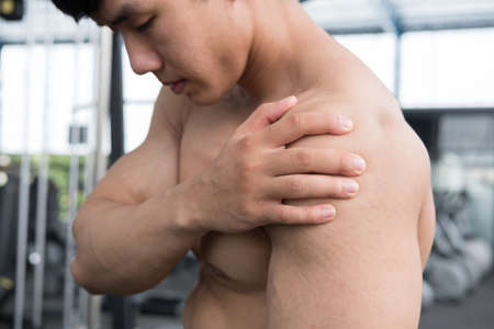 muscular male having pain on shoulder in gym.  young man injure while workout in fitness center. asian bodybuilder feel hurt while exercise at health club. Sports injury, muscle intense.の写真素材