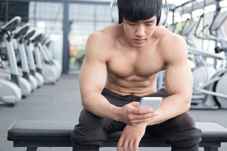 young man use mobile phone in fitness center. male athlete listen to music in cellphone in gym. sporty asian guy resting in health club after working out.の写真素材