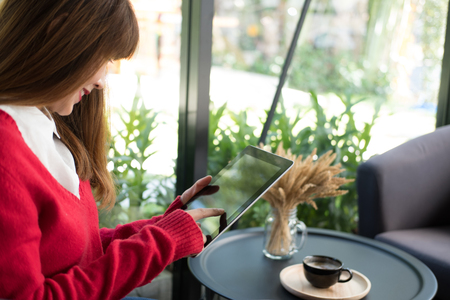 young woman wearing red sweater typing text message on application on tablet at home. female adult sitting on armchair using touchpad for social networking at cafe. technology, lifestyle, peopleの写真素材
